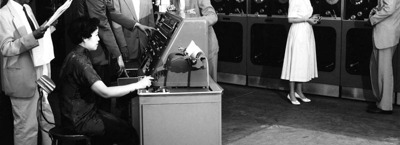 Man in the forground reasds a newpaper as a womena opperates at terminal. Three other men look over her shoulder. Grace Hopper discusses with a man in the backgroun